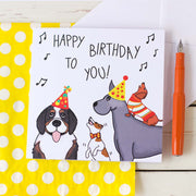 Birthday card illustrated by fizzijayne. A Bernese Mountain Dog wearing a party hat, a Jack Russell wearing a Bow tie jumping up to get to the height of the big dogs, a Great Dane wearing a party hat with a Dachshund on it's back wearing a stripey jumper. Image is with a white envelope and spotty paper bag that cards are packaged in